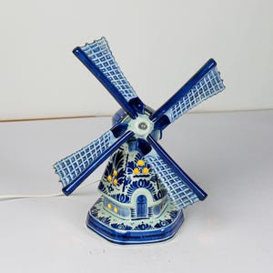 Peut inclure: Lampe en céramique en forme de moulin à vent, bleue et blanche. Les pales sont bleu foncé avec un motif en treillis. La base présente un motif floral détaillé et de petites fenêtres qui émettent une lueur chaude. Un cordon blanc sort de la base.