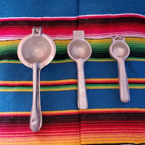 May include: Three silver lime squeezers on a colorful striped fabric background. The largest squeezer has a handle with the words "Made in Mexico" printed on it. The two smaller squeezers have handles with the words "Made in USA" printed on them.