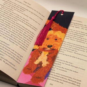 Può includere: Un segnalibro con l'illustrazione di un cane marrone-arancione e un nappa rosa. Il segnalibro è inserito in un libro aperto, con testo visibile sulle pagine.