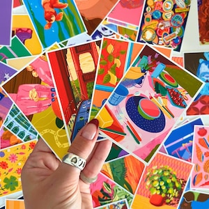 Puede incluir: Una mano sosteniendo una colección de coloridas impresiones artísticas. Las impresiones presentan varias ilustraciones, incluyendo un puente, limones y una mesa puesta. Las impresiones son en tonos de rojo, naranja, azul y rosa. Se ve un anillo plateado con recortes de corazón.