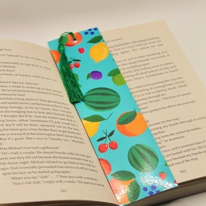 Puede incluir: Un marcapáginas colorido con un fondo turquesa y un patrón de frutas, incluyendo sandías, naranjas, limones, ciruelas y cerezas. Una borla verde cuelga de la parte superior. El marcapáginas está en un libro abierto.