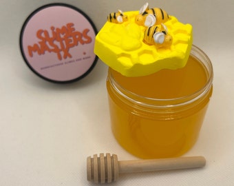 May include: A clear glass jar filled with yellow honey, topped with a yellow honeycomb lid with small bee figurines. A wooden honey dipper is included.