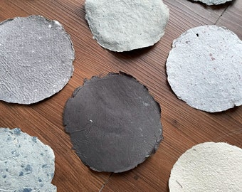 May include: Assortment of round, handmade paper discs in various shades of blue, gray, white, and black. The paper has a textured, organic look.