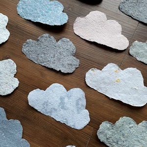 Könnte beinhalten: Eine Sammlung handgemachter Papierwolken in verschiedenen Farben und Texturen, darunter Grau, Blau, Rosa und Weiß. Die Wolken sind auf einer Holzoberfläche angeordnet.