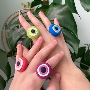 Puede incluir: Cuatro anillos de ojo turco coloridos hechos de arcilla polimérica. Los anillos son en tonos de verde, azul, rosa y morado. Cada anillo tiene un círculo blanco alrededor del ojo y una pupila negra.