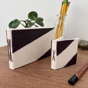 May include: Two handmade fabric wallets with a geometric design. The wallets are cream-colored with a diagonal dark burgundy stripe. The larger wallet has the letter "M" stitched on the side, and the smaller wallet has the letter "X".