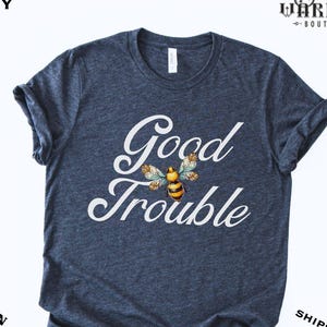 May include: Heather navy t-shirt with the words "Good Trouble" in white script, featuring a bee illustration. The shirt is a dark blue color and has a crew neck. The text "Veteran Owned" is also visible.