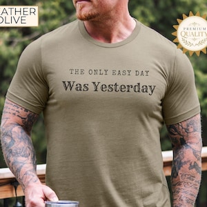 Könnte beinhalten: Ein kurzärmeliges T-Shirt in Heather Olive mit dem schwarzen Schriftzug "THE ONLY EASY DAY WAS YESTERDAY" auf der Brust. Das Shirt hat einen Rundhalsausschnitt und eine Standardpassform und zeigt ein einfarbiges Design ohne weitere Muster.