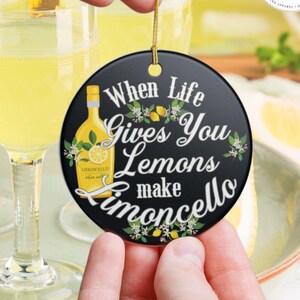 May include: A round black ceramic ornament featuring a yellow bottle illustration and the white text "When Life Gives You Lemons make Limoncello," accented with small lemon and blossom graphics, suspended by a thin gold cord for hanging.
