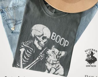 May include: A dark gray t-shirt with a skeleton and corgi graphic, the word "BOOP" is printed on the shirt. The shirt is paired with light blue jeans and a tan hat. The text "comfort colors" and "pepper" are printed on the bottom.
