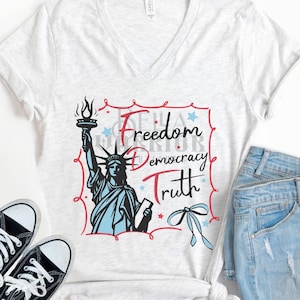 May include: A light gray V-neck t-shirt with a graphic of the Statue of Liberty holding a torch, with the words "Freedom, Democracy, Truth." The shirt is paired with denim shorts and black sneakers.