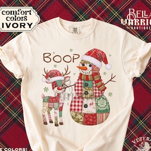 May include: Ivory-colored t-shirt featuring a patchwork reindeer and snowman design with the word "BOOP". The snowman wears a red Santa hat and scarf. The reindeer also wears a Santa hat. The shirt has the words "Comfort Colors" and "Bella Warrior Boutique" printed on it.