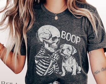 May include: Dark grey heather t-shirt featuring a skeleton and a bulldog with the word "BOOP" above them. The skeleton is holding a bone up to the dog's nose. The shirt has short sleeves and a crew neck.
