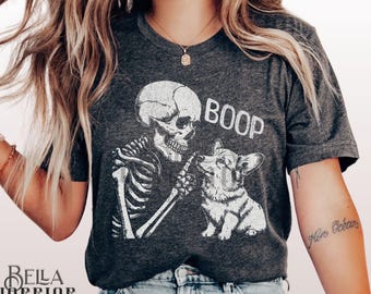 May include: A dark grey heather t-shirt featuring a white graphic of a skeleton touching the nose of a corgi dog. The text BOOP is printed in white above the dog. The shirt has short sleeves and a crew neckline, presented in a casual style.