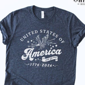 Puede incluir: Camiseta azul marino jaspeada con un dise&ntilde;o blanco que dice "United States of America" con un &aacute;guila y estrellas. El texto "250 Years 1776-2026" tambi&eacute;n est&aacute; impreso. De Bella Warrior Boutique.
