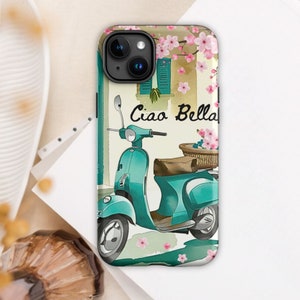 Op de afbeelding: Een turquoise blauwe Vespa scooter met een mand vol bloemen achterop. De scooter staat geparkeerd voor een gebouw met een roze bloeiende struik en de tekst "Ciao Bella" aan de zijkant. De achtergrond is een wit-lichtblauw bloemenpatroon.
