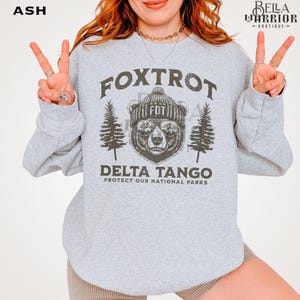 Puede incluir: Una sudadera gris con un oso negro que lleva un gorro que dice "FDT" y gafas de sol. El texto "Fox Trot Delta Tango Protect Our National Parks" est&aacute; impreso en la sudadera.