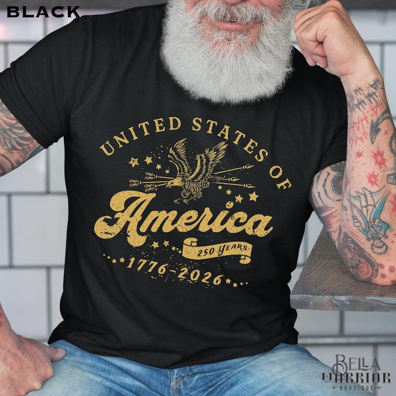 Puede incluir: Camiseta negra con un dise&ntilde;o dorado que dice "United States of America" con un &aacute;guila y flechas. El texto "1776-2026" y "250 Years" tambi&eacute;n est&aacute;n incluidos en el dise&ntilde;o. La camiseta es de cuello redondo.
