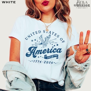 Puede incluir: Camiseta blanca con un gr&aacute;fico azul que dice "United States of America" con un &aacute;guila y los a&ntilde;os "1776-2026". La camiseta tiene cuello redondo y mangas cortas. El dise&ntilde;o tambi&eacute;n incluye el texto "250 Years".