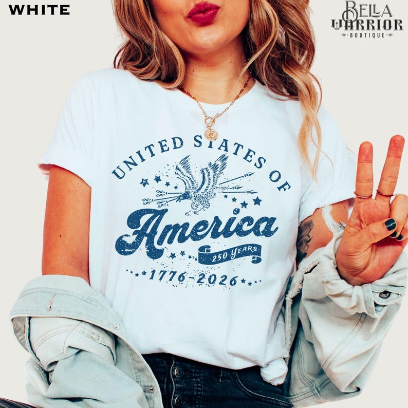 Puede incluir: Camiseta blanca con un gr&aacute;fico azul que dice "United States of America" con un &aacute;guila y los a&ntilde;os "1776-2026". La camiseta tiene cuello redondo y mangas cortas. El dise&ntilde;o tambi&eacute;n incluye el texto "250 Years".