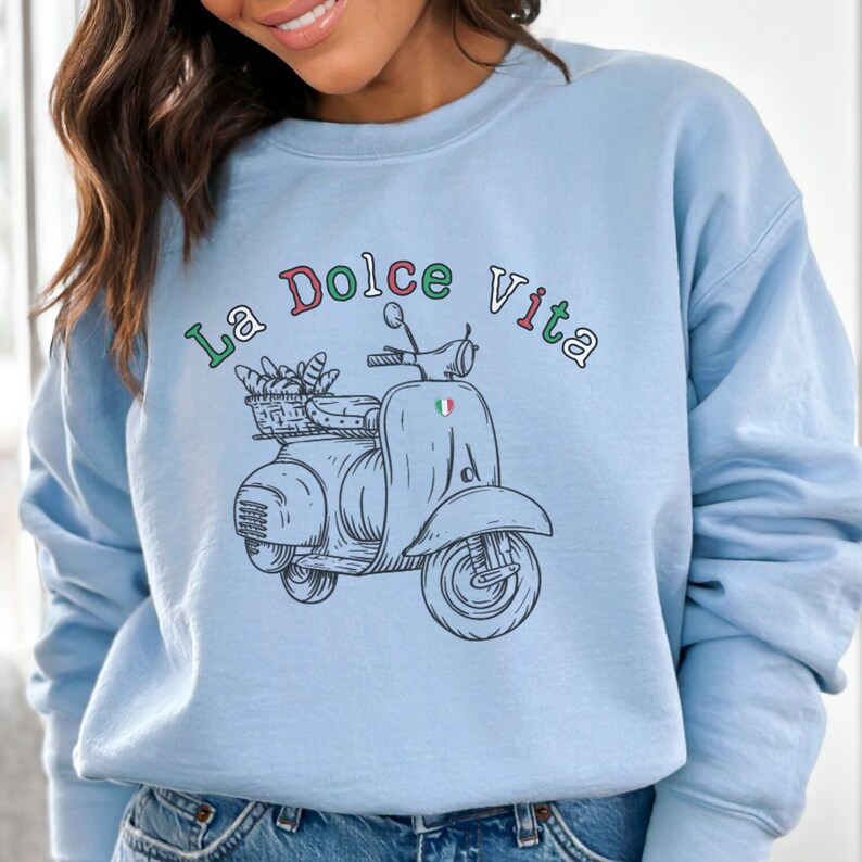 May include: A light blue sweatshirt featuring a graphic of a vintage scooter carrying a basket of bread. The text "La Dolce Vita" is written in a colourful arc above the scooter.