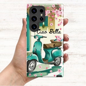 Op de afbeelding: Een turquoise blauwe scooter met een rieten mand gevuld met roze bloemen. De scooter staat geparkeerd voor een gebouw met een raam en roze bloemen aan een wijnstok. De tekst "Ciao Bella" staat op de afbeelding.