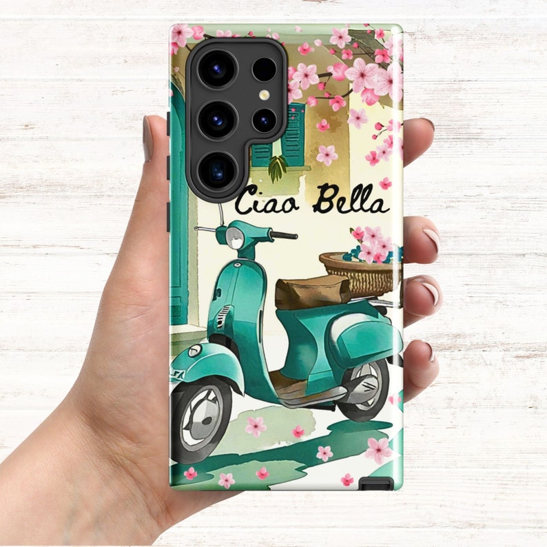 Op de afbeelding: Een turquoise blauwe scooter met een rieten mand gevuld met roze bloemen. De scooter staat geparkeerd voor een gebouw met een raam en roze bloemen aan een wijnstok. De tekst "Ciao Bella" staat op de afbeelding.