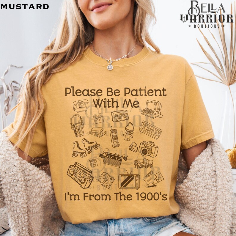 Puede incluir: Una camiseta de manga corta color amarillo mostaza con ilustraciones de l&iacute;nea marr&oacute;n de objetos de los a&ntilde;os 1900, incluyendo un reproductor de casetes, una c&aacute;mara y una computadora retro. El texto dice: Please Be Patient With Me I'm From The 1900's.