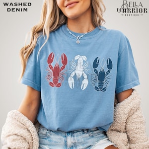 May include: Light blue t-shirt with three lobster illustrations in red, white, and navy blue. The shirt has the words "Washed Denim" in the upper left corner and "Bella Warrior Boutique" in the upper right corner.