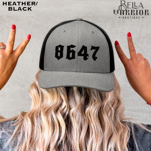 May include: Heather and black trucker hat with the number "8647" in bold, black lettering. The hat has a gray front panel and a black mesh back. The text "HEATHER/BLACK" is in the upper left corner. The Bella Warrior Boutique logo is in the upper right corner.
