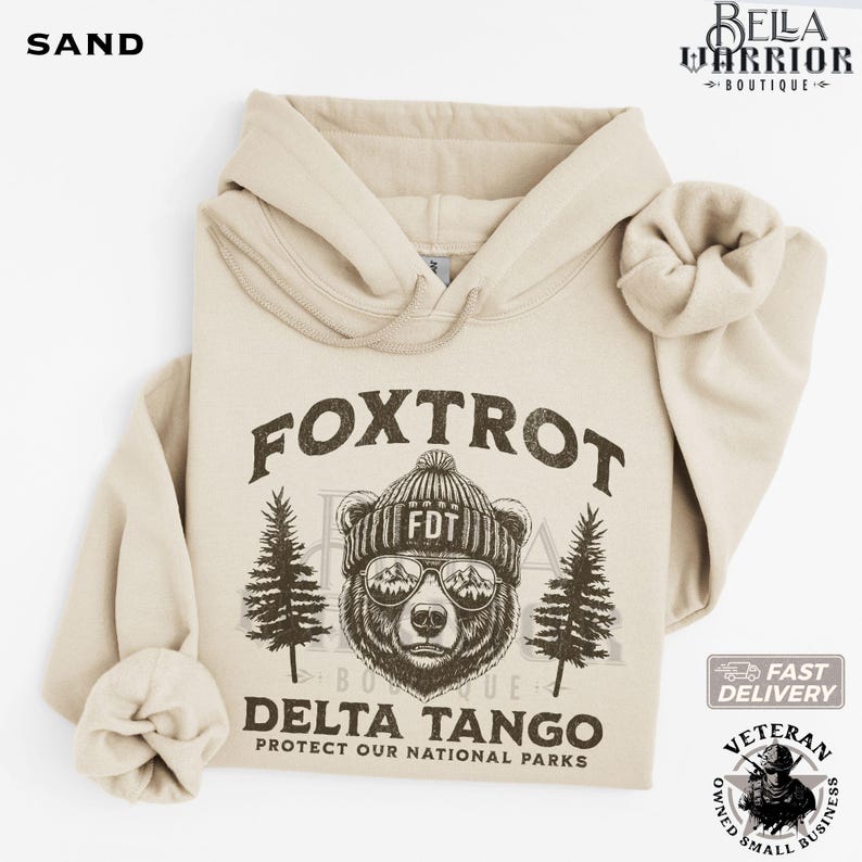 Puede incluir: Una sudadera con capucha beige con un oso marr&oacute;n que lleva un sombrero y gafas de sol. El texto "FOXTROT DELTA TANGO PROTECT OUR NATIONAL PARKS" est&aacute; impreso en la sudadera.