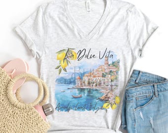 May include: A light grey v-neck t-shirt with a watercolor illustration of a coastal Italian town with the text "La Dolce Vita Amalfi Coast Italy".