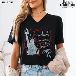 May include: Black V-neck t-shirt featuring a graphic of the Statue of Liberty holding a torch, with the words "Freedom Democracy Truth" in red and white. The design includes stars and a bow.