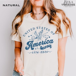 Puede incluir: Camiseta color crema con un gr&aacute;fico azul desgastado que dice "United States of America" con una ilustraci&oacute;n de un &aacute;guila. Tambi&eacute;n incluye el texto "1776-2026". La camiseta es de color natural.