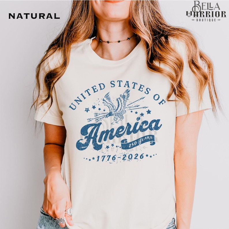 Puede incluir: Camiseta color crema con un gr&aacute;fico azul desgastado que dice "United States of America" con una ilustraci&oacute;n de un &aacute;guila. Tambi&eacute;n incluye el texto "1776-2026". La camiseta es de color natural.