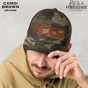 May include: A camouflage trucker hat with a brown mesh back and a curved brim. The front panel features an orange design. The hat has the text "CAMO/ BROWN ORANGE" and the logo "BELLA WARRIOR BOUTIQUE".
