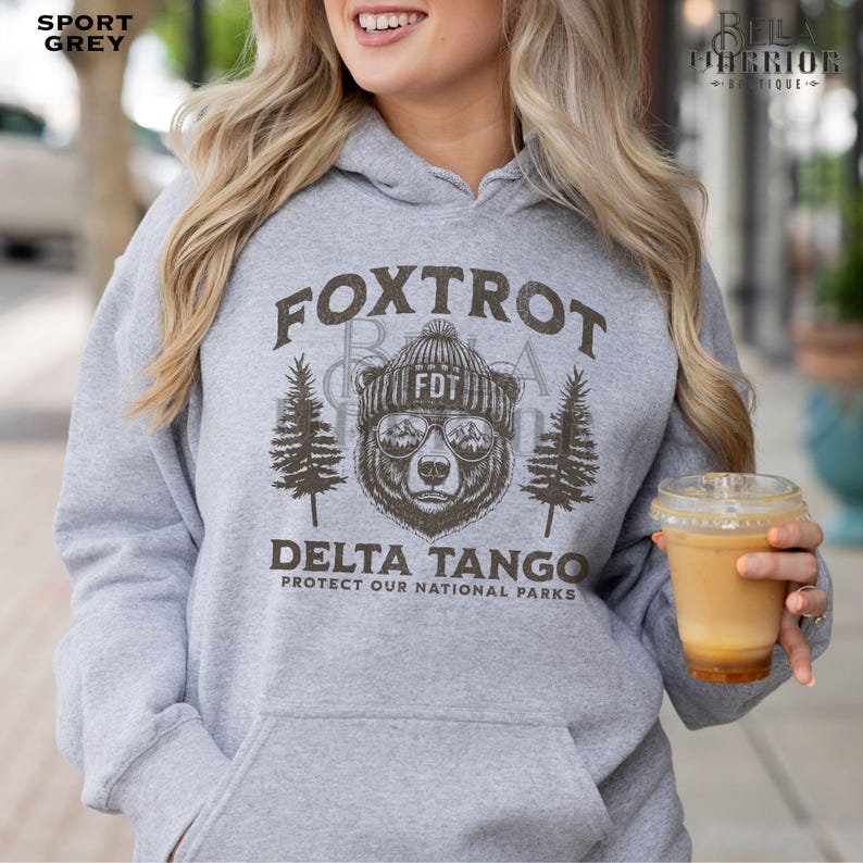 Puede incluir: Una sudadera con capucha gris con un estampado gr&aacute;fico de un oso con un gorro y gafas de sol. El texto "Fox Trot Delta Tango Protect Our National Parks" est&aacute; impreso en la sudadera.