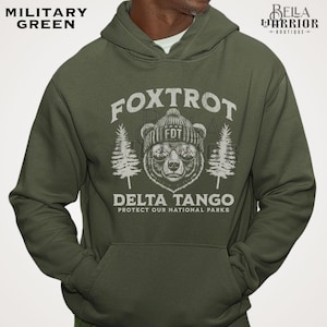 Puede incluir: Una sudadera con capucha verde con un gr&aacute;fico blanco de un oso con un sombrero y gafas de sol. El texto "Fox Trot Delta Tango Protect Our National Parks" est&aacute; impreso en la sudadera.
