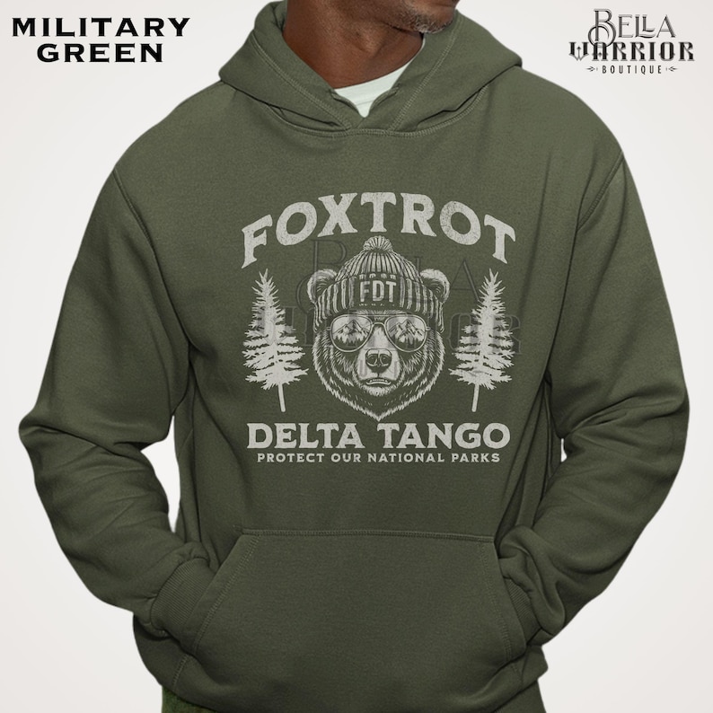 Puede incluir: Una sudadera con capucha verde con un gr&aacute;fico blanco de un oso con un sombrero y gafas de sol. El texto "Fox Trot Delta Tango Protect Our National Parks" est&aacute; impreso en la sudadera.
