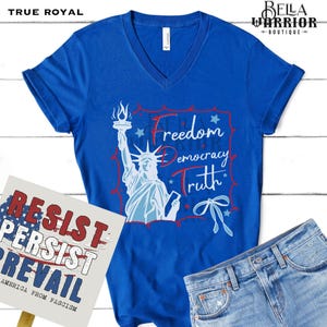 May include: A blue V-neck t-shirt with a graphic of the Statue of Liberty and the words "Freedom, Democracy, Truth." A sign with the words "Resist, Persist, Prevail" is in the image. The shirt is paired with denim jeans.