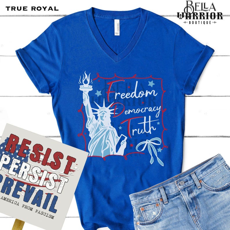 May include: A blue V-neck t-shirt with a graphic of the Statue of Liberty and the words "Freedom, Democracy, Truth." A sign with the words "Resist, Persist, Prevail" is in the image. The shirt is paired with denim jeans.
