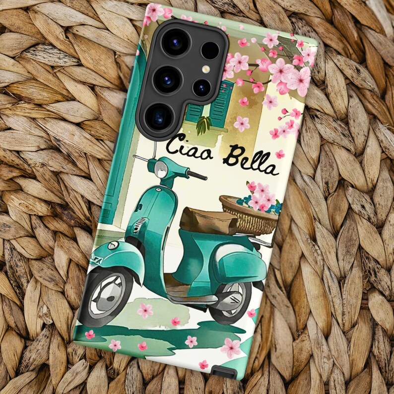 Op de afbeelding: Een turquoise scooter met een mand vol bloemen, een wit gebouw met een raam en de tekst "Ciao Bella" op een witte achtergrond met roze bloemen.