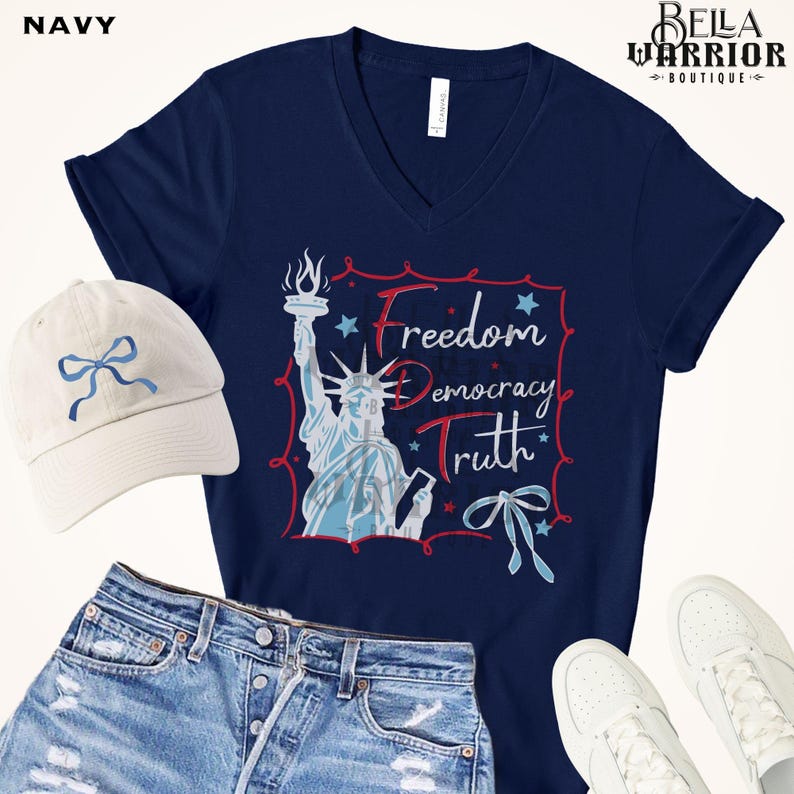 May include: Navy blue V-neck t-shirt with a graphic of the Statue of Liberty and the words "Freedom, Democracy, Truth." A white baseball cap with a blue bow and denim shorts are also in the image. White sneakers complete the patriotic theme.