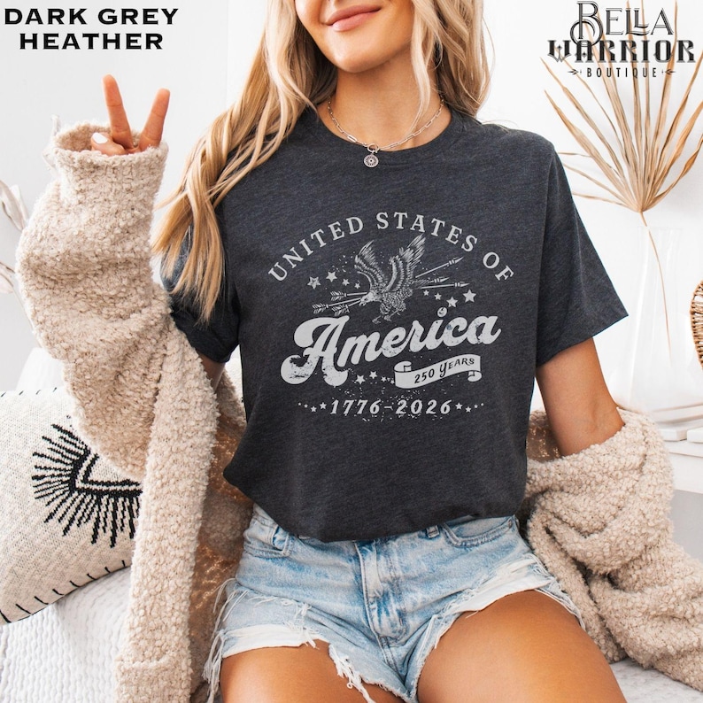 Puede incluir: Camiseta gris oscuro jaspeado con el texto "United States of America" y un gr&aacute;fico de &aacute;guila. La camiseta tambi&eacute;n presenta el texto "250 Years" y "1776-2026".