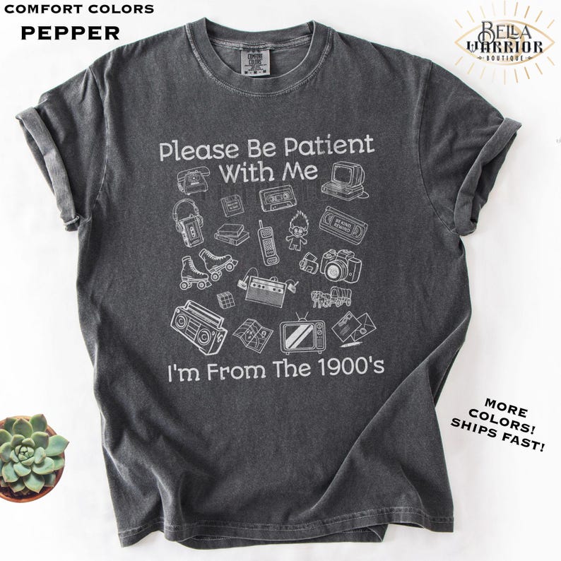 Puede incluir: Una camiseta gris carb&oacute;n de manga corta con ilustraciones en l&iacute;nea blanca de objetos vintage que incluyen un reproductor de casetes, patines y un radiocasete. El texto dice: Please Be Patient With Me I'm From The 1900's. Tiene cuello redondo.