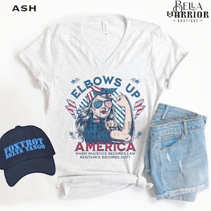Puede incluir: Una camiseta blanca de cuello en V con un gráfico de una mujer con cabello rojo, blanco y azul y gafas de sol. El gráfico dice "Elbows Up America" y "When injustice becomes law, resistance becomes duty".