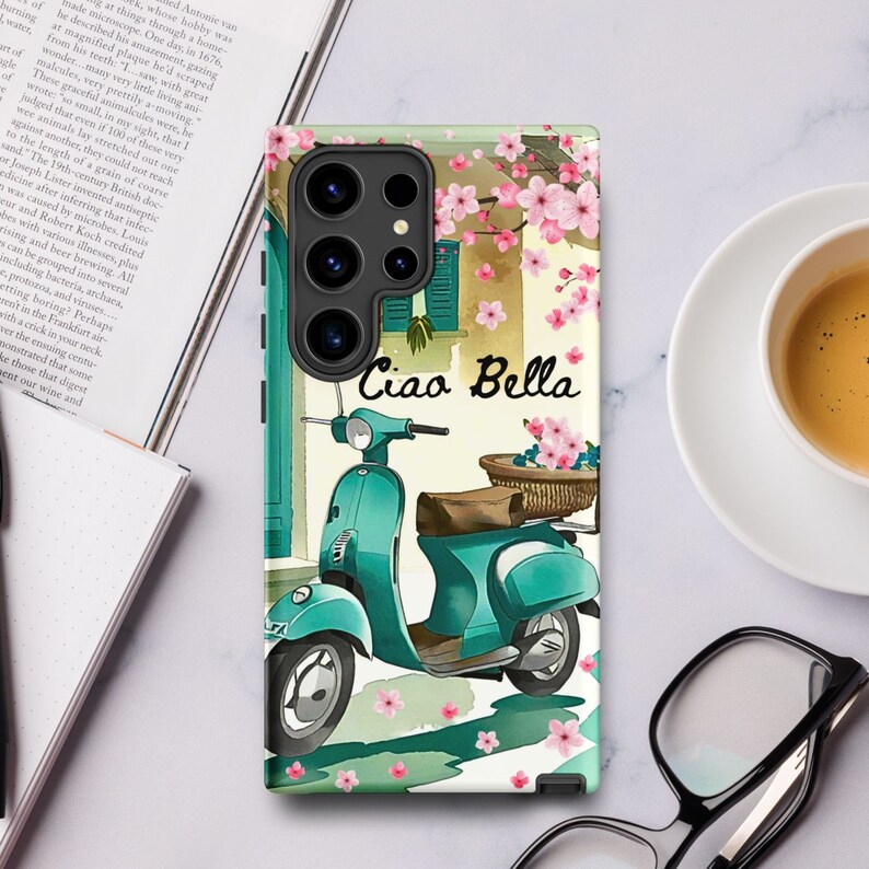 Op de afbeelding: Een turquoise blauwe scooter met een mand vol roze bloemen achterop, geparkeerd voor een gebouw met roze bloemen aan een wijnstok. De tekst "Ciao Bella" staat in zwart op een witte achtergrond.