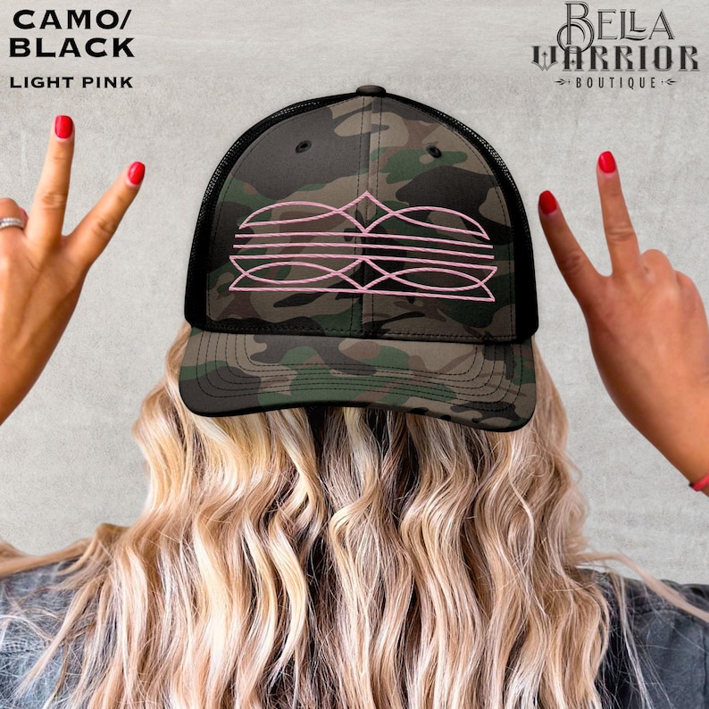 May include: Camo and black trucker hat with a pink graphic design. The hat has a mesh back and a curved brim. The text "CAMO/BLACK" and "LIGHT PINK" is visible in the upper left corner. The Bella Warrior Boutique logo is in the upper right corner.