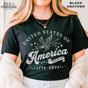 Puede incluir: Camiseta negra jaspeada con un dise&ntilde;o gr&aacute;fico desgastado. El dise&ntilde;o presenta el texto "United States of America" alrededor de un &aacute;guila con flechas, estrellas y "America 250 Years 1776-2026". El logotipo de Bella Warrior Boutique est&aacute; en la esquina superior izquierda.