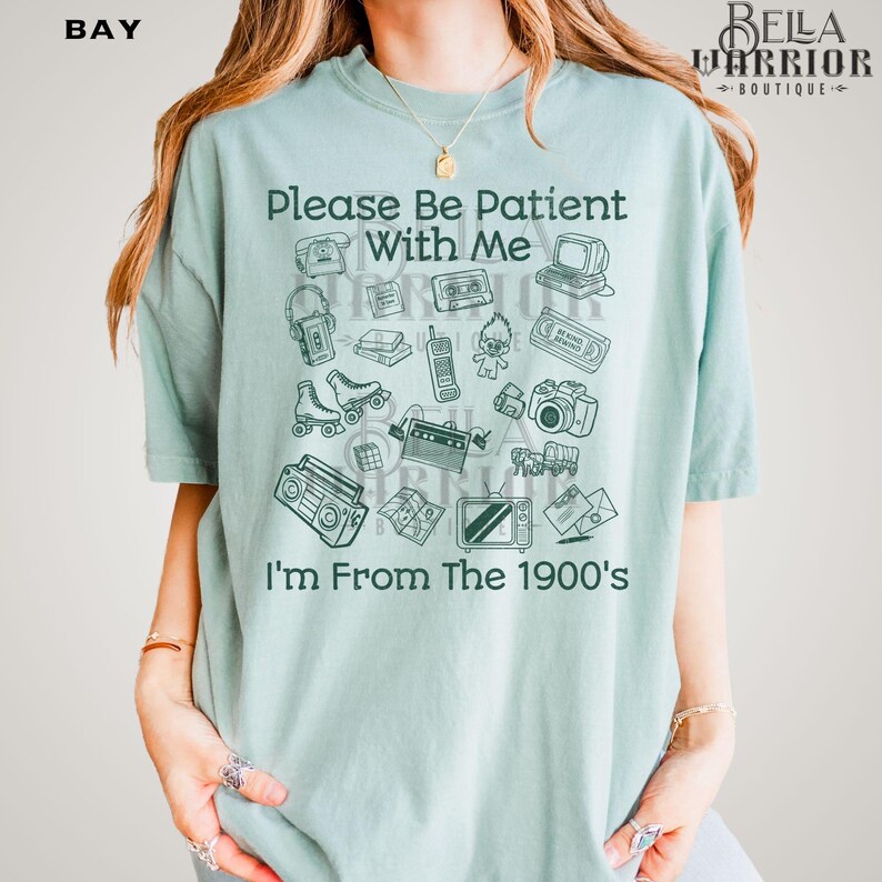 Puede incluir: Una camiseta de manga corta de color verde claro con texto en verde oscuro que dice Please Be Patient With Me I'm From The 1900's. El gr&aacute;fico incluye ilustraciones de objetos retro como un reproductor de casetes, patines y una c&aacute;mara.